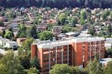A dense urban neighborhood with numerous brick buildings surrounded by lush green trees. The foreground features a large, multi-story brick building, while the background is filled with a mix of residential homes and abundant greenery. The area appears orderly, with well-maintained streets and a few visible utility poles.