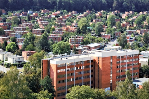 A dense urban neighborhood with numerous brick buildings surrounded by lush green trees. The foreground features a large, multi-story brick building, while the background is filled with a mix of residential homes and abundant greenery. The area appears orderly, with well-maintained streets and a few visible utility poles.