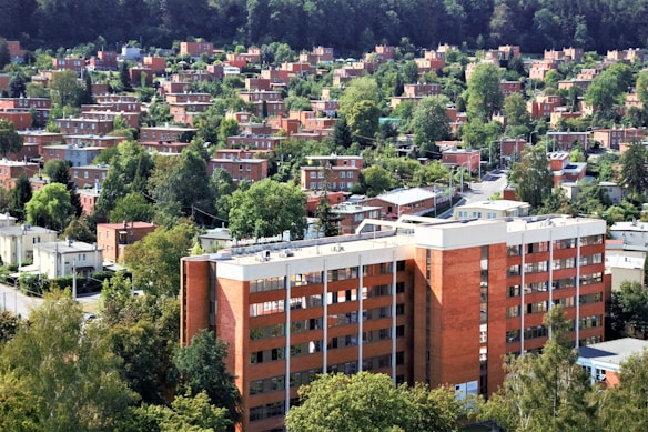A dense urban neighborhood with numerous brick buildings surrounded by lush green trees. The foreground features a large, multi-story brick building, while the background is filled with a mix of residential homes and abundant greenery. The area appears orderly, with well-maintained streets and a few visible utility poles.