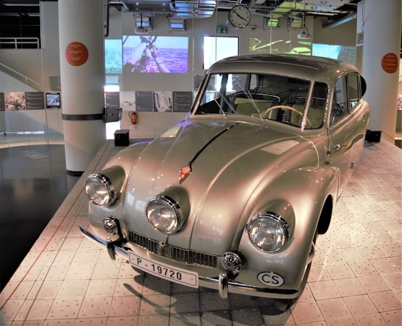 A vintage car with a glossy grey exterior is displayed in a modern museum setting. The car features four prominent round headlights and a unique aerodynamic design. A screen with historical videos is seen in the background, setting a nostalgic and educational atmosphere. The museum space is well-lit with various exhibits and informative plaques.