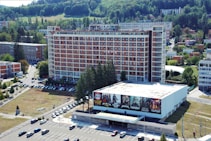Aerial view of a large multi-story hotel building with a red and white facade and many windows. In front of the hotel, there is a smaller building with large promotional banners featuring various bands or events. A parking lot with multiple cars is visible, along with several trees and grassy areas. The surrounding area includes more buildings and green spaces, with a forested hillside in the background.