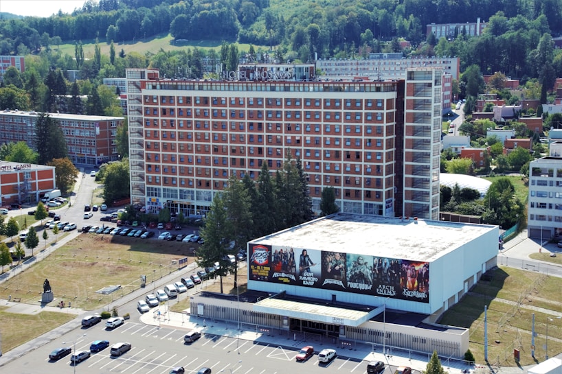 Aerial view of a large multi-story hotel building with a red and white facade and many windows. In front of the hotel, there is a smaller building with large promotional banners featuring various bands or events. A parking lot with multiple cars is visible, along with several trees and grassy areas. The surrounding area includes more buildings and green spaces, with a forested hillside in the background.