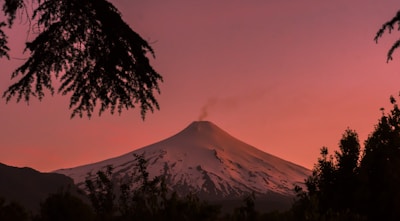 snow-covered, smoking volcano top seen from a forest at its foot