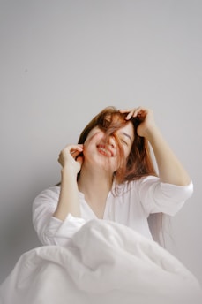 A person is smiling joyfully, with their head tilted slightly back and hair playfully swept over their face. They are wearing a white shirt and are partially covered with a white blanket.