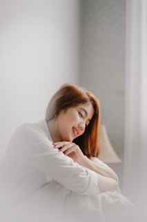 A person with long hair is sitting with their eyes closed and a gentle smile, wearing a light-colored shirt. Their hands are clasped together, resting on their chest, giving a serene and peaceful appearance. In the background, there is a soft-focus lamp, adding to the cozy atmosphere.