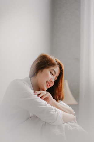 A person with long hair is sitting with their eyes closed and a gentle smile, wearing a light-colored shirt. Their hands are clasped together, resting on their chest, giving a serene and peaceful appearance. In the background, there is a soft-focus lamp, adding to the cozy atmosphere.