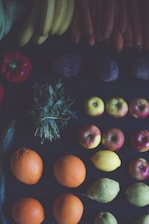 A variety of fruits and vegetables are artfully arranged in rows. Brightly colored oranges, apples, and lemons are positioned alongside bananas, carrots, and peppers. Green leafy herbs and other vegetables like pears and potatoes complete the arrangement on a dark background.