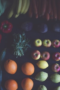 A variety of fruits and vegetables are artfully arranged in rows. Brightly colored oranges, apples, and lemons are positioned alongside bananas, carrots, and peppers. Green leafy herbs and other vegetables like pears and potatoes complete the arrangement on a dark background.