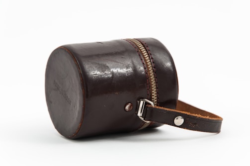 A small, cylindrical leather case with a zipper running along the middle and a strap attached to the side. The leather is dark brown, slightly glossy, and appears to be well-crafted.