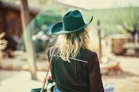 A person with long hair is wearing a dark cowboy hat and a black shirt, standing in an outdoor, rustic setting. The background is blurred, featuring earthy tones and wooden structures, giving a sense of being in a rural area.