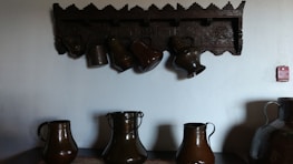 Several dark ceramic jugs are placed on a surface, with a few hanging from an intricately carved wooden shelf above them on a plain wall. The jugs have a glossy finish, and the wall decoration has a geometric pattern. There is a small red sign to the right on the wall.
