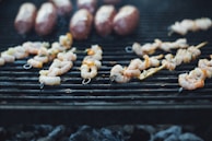 Shrimp skewers and several sausages are grilling over a charcoal barbecue. The skewers are neatly arranged, showcasing their grilled texture, with bits of seasoning visible. The metal grill grates are blackened from use, and there is a soft focus on the smoky background.