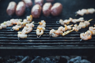 Shrimp skewers and several sausages are grilling over a charcoal barbecue. The skewers are neatly arranged, showcasing their grilled texture, with bits of seasoning visible. The metal grill grates are blackened from use, and there is a soft focus on the smoky background.
