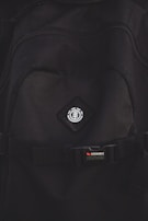 A close-up view of a black backpack with a logo featuring a tree icon inside a circle, placed on a diamond-shaped patch. Below the logo, there is a strap with a buckle bearing the text 'STOWABLE SKATEBOARD FRONT STRAPS' in white and red letters.