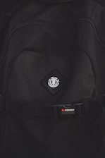 A close-up view of a black backpack with a logo featuring a tree icon inside a circle, placed on a diamond-shaped patch. Below the logo, there is a strap with a buckle bearing the text 'STOWABLE SKATEBOARD FRONT STRAPS' in white and red letters.
