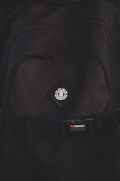 A close-up view of a black backpack with a logo featuring a tree icon inside a circle, placed on a diamond-shaped patch. Below the logo, there is a strap with a buckle bearing the text 'STOWABLE SKATEBOARD FRONT STRAPS' in white and red letters.