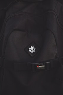 A close-up view of a black backpack with a logo featuring a tree icon inside a circle, placed on a diamond-shaped patch. Below the logo, there is a strap with a buckle bearing the text 'STOWABLE SKATEBOARD FRONT STRAPS' in white and red letters.