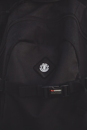 A close-up view of a black backpack with a logo featuring a tree icon inside a circle, placed on a diamond-shaped patch. Below the logo, there is a strap with a buckle bearing the text 'STOWABLE SKATEBOARD FRONT STRAPS' in white and red letters.