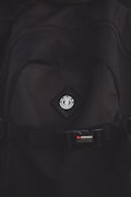 A close-up view of a black backpack with a logo featuring a tree icon inside a circle, placed on a diamond-shaped patch. Below the logo, there is a strap with a buckle bearing the text 'STOWABLE SKATEBOARD FRONT STRAPS' in white and red letters.