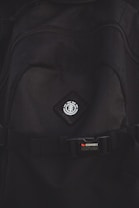 A close-up view of a black backpack with a logo featuring a tree icon inside a circle, placed on a diamond-shaped patch. Below the logo, there is a strap with a buckle bearing the text 'STOWABLE SKATEBOARD FRONT STRAPS' in white and red letters.