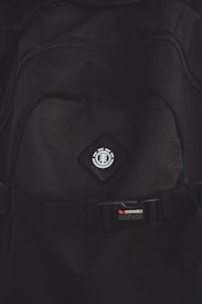 A close-up view of a black backpack with a logo featuring a tree icon inside a circle, placed on a diamond-shaped patch. Below the logo, there is a strap with a buckle bearing the text 'STOWABLE SKATEBOARD FRONT STRAPS' in white and red letters.