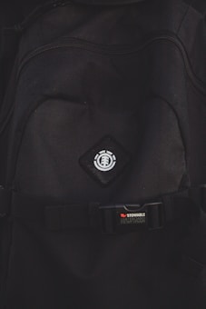 A close-up view of a black backpack with a logo featuring a tree icon inside a circle, placed on a diamond-shaped patch. Below the logo, there is a strap with a buckle bearing the text 'STOWABLE SKATEBOARD FRONT STRAPS' in white and red letters.