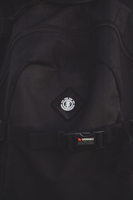 A close-up view of a black backpack with a logo featuring a tree icon inside a circle, placed on a diamond-shaped patch. Below the logo, there is a strap with a buckle bearing the text 'STOWABLE SKATEBOARD FRONT STRAPS' in white and red letters.