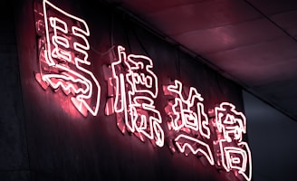 Bright pink neon Chinese characters illuminated against a dark wall, creating a glowing effect. The light reflects softly on the surrounding surfaces, emphasizing the vibrant lettering.