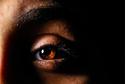 A close-up view of a human eye, with rich brown iris, surrounded by shadows. The lighting emphasizes the texture and details of the skin around the eye, with the eyebrow visible at the top and eyelashes clearly defined against the darkness.