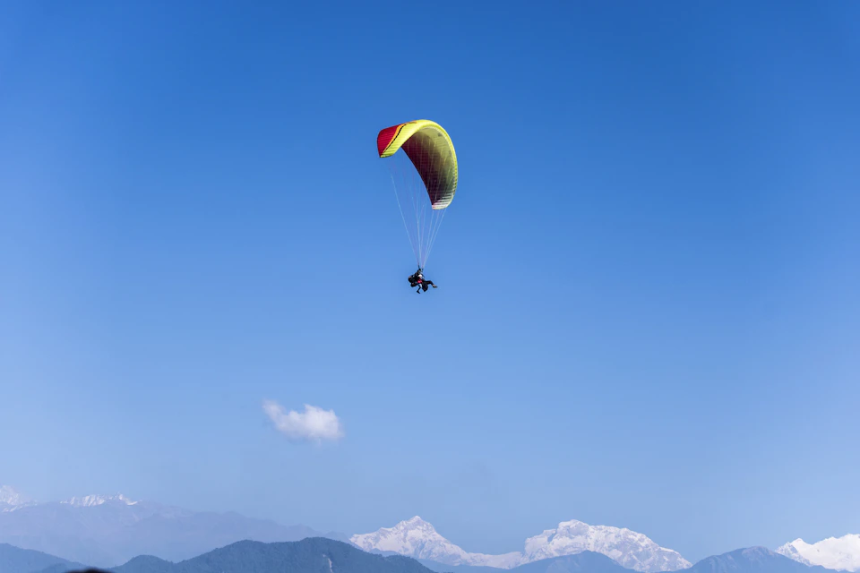 Person paragliding high above mountain range under clear blue sky with fluffy white clouds