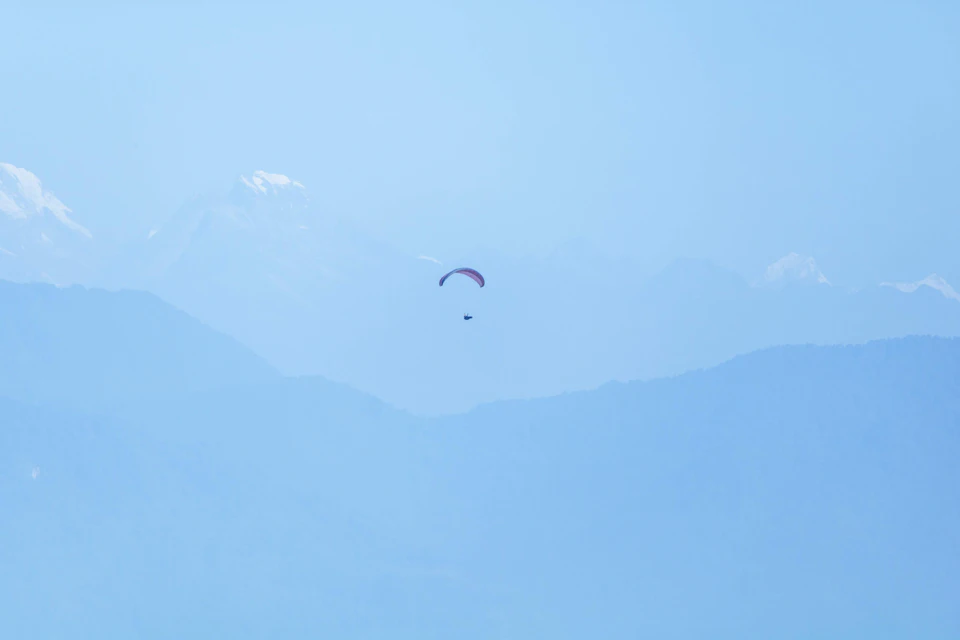 The image depicts a paraglider soaring high above a mountainous landscape, with their parachute trailing behind them against a clear blue sky. The mountains are covered in lush green trees and snow-capped peaks, creating a breathtaking backdrop for the paraglider's journey.
