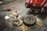A red classic car is parked in a garage with its trunk open. Nearby, various tools and a spare tire are laid out on the floor, including jumper cables, a tire iron, and a toolbox. The atmosphere suggests a repair or maintenance activity in progress.
