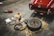 A red classic car is parked in a garage with its trunk open. Nearby, various tools and a spare tire are laid out on the floor, including jumper cables, a tire iron, and a toolbox. The atmosphere suggests a repair or maintenance activity in progress.
