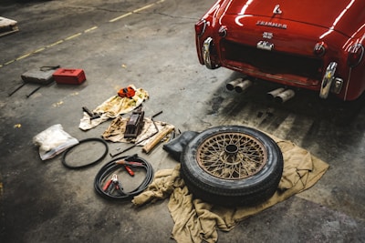 A red classic car is parked in a garage with its trunk open. Nearby, various tools and a spare tire are laid out on the floor, including jumper cables, a tire iron, and a toolbox. The atmosphere suggests a repair or maintenance activity in progress.