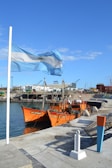 A coastal scene with a gently waving light blue and white flag beside a vibrant orange fishing boat docked at the port. The background features industrial buildings and other maritime equipment against a clear blue sky.