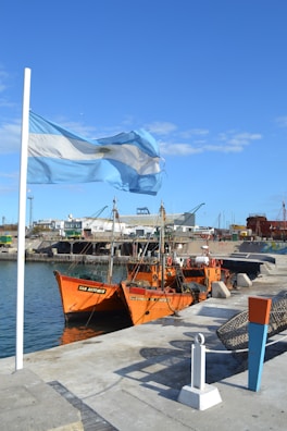 A coastal scene with a gently waving light blue and white flag beside a vibrant orange fishing boat docked at the port. The background features industrial buildings and other maritime equipment against a clear blue sky.