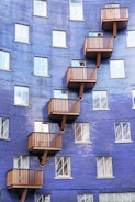A building facade covered in blue tiles features multiple wooden balconies arranged in a zig-zag pattern. The structure includes numerous rectangular windows with white frames, contributing to a geometric and modern aesthetic.