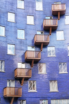 A building facade covered in blue tiles features multiple wooden balconies arranged in a zig-zag pattern. The structure includes numerous rectangular windows with white frames, contributing to a geometric and modern aesthetic.