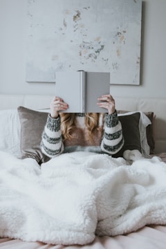 A person is comfortably lying in bed, covered with a fluffy white blanket, and holding an open book in front of their face. They are wearing a cozy striped sweater. The background features a light abstract painting on the wall, adding to the peaceful ambiance.