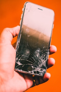 A hand is holding a smartphone with a severely cracked screen against an orange background. The phone is damaged, with visible cracks running across its surface.