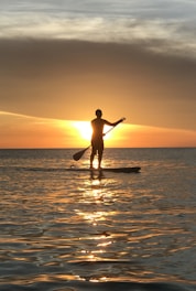 A person is silhouetted against a vibrant sunset while standing on a paddleboard in calm waters. The water reflects the warm tones of the sunset, creating a serene and picturesque scene.