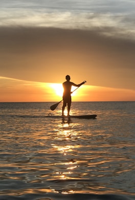 A person is silhouetted against a vibrant sunset while standing on a paddleboard in calm waters. The water reflects the warm tones of the sunset, creating a serene and picturesque scene.