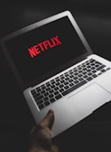 A hand is holding an open laptop displaying the Netflix logo on the screen. The image is taken in a dimly lit environment, highlighting the laptop's keyboard and screen.