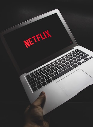 A hand is holding an open laptop displaying the Netflix logo on the screen. The image is taken in a dimly lit environment, highlighting the laptop's keyboard and screen.