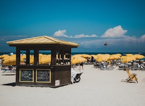 A sunny beach scene featuring numerous yellow umbrellas and lounge chairs arranged on the sand. A small wooden hut with signs is centrally located, offering rental items. The ocean is visible in the background with a person parasailing in the distance under a partly cloudy sky.