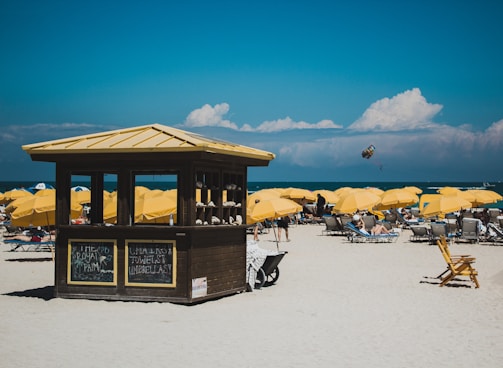 A sunny beach scene featuring numerous yellow umbrellas and lounge chairs arranged on the sand. A small wooden hut with signs is centrally located, offering rental items. The ocean is visible in the background with a person parasailing in the distance under a partly cloudy sky.