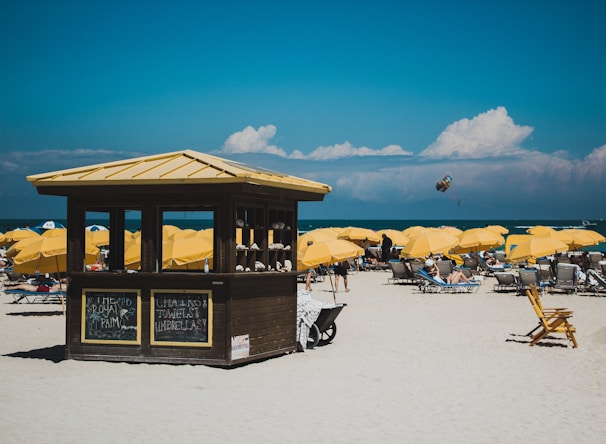 A sunny beach scene featuring numerous yellow umbrellas and lounge chairs arranged on the sand. A small wooden hut with signs is centrally located, offering rental items. The ocean is visible in the background with a person parasailing in the distance under a partly cloudy sky.