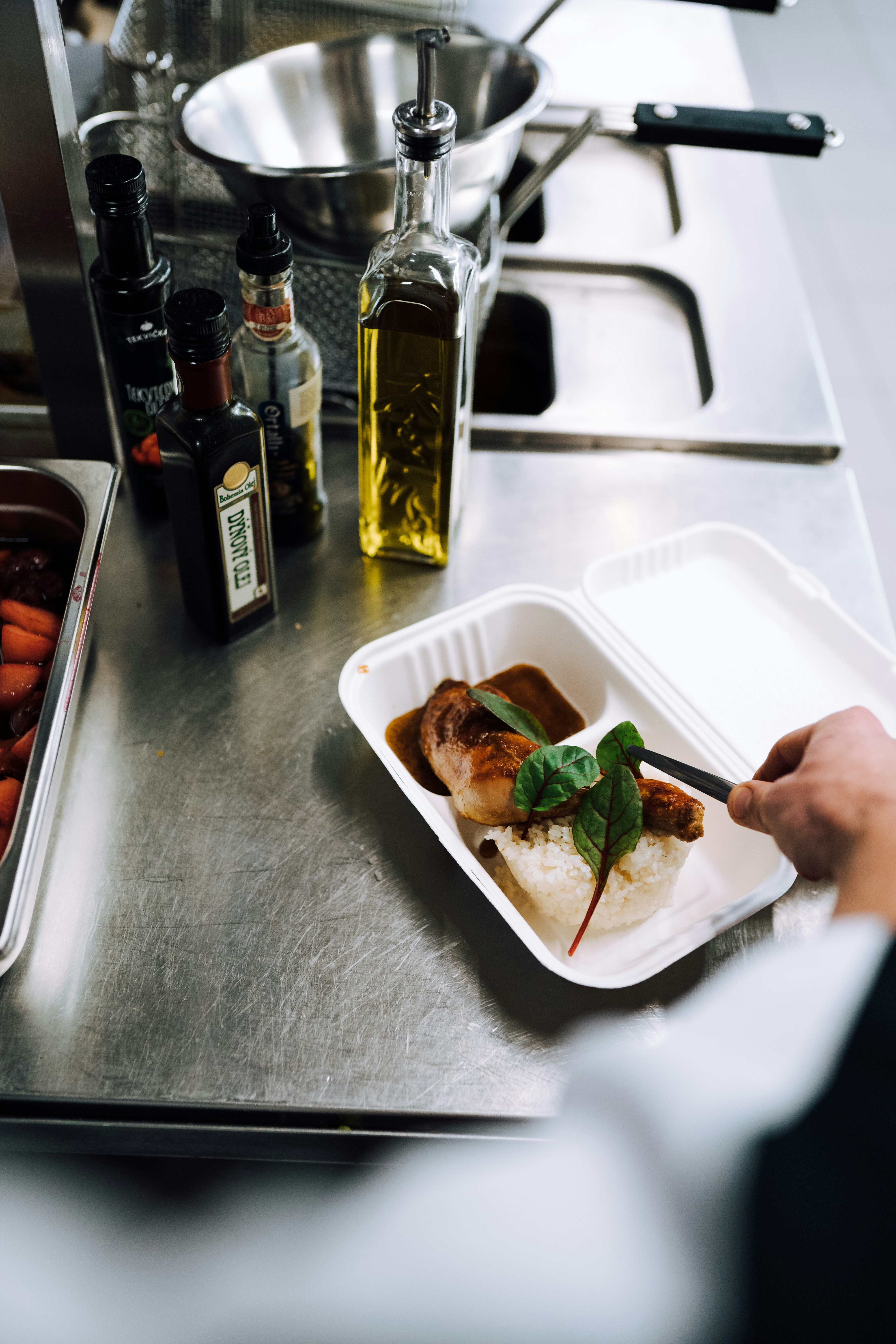 On a stainless steel counter, a hand is placing a prepared meal into a white takeout container. The meal consists of rice, a piece of meat with sauce, and a garnish of green leafy vegetable. Adjacent to the container, there are bottles of olive oil and various vinegars. A metal mixing bowl and kitchen utensils can be seen in the background.