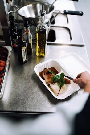 On a stainless steel counter, a hand is placing a prepared meal into a white takeout container. The meal consists of rice, a piece of meat with sauce, and a garnish of green leafy vegetable. Adjacent to the container, there are bottles of olive oil and various vinegars. A metal mixing bowl and kitchen utensils can be seen in the background.