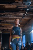 A woman is wearing a matching athletic outfit with a tropical leaf pattern, featuring a crop top and leggings. She is walking down what appears to be an urban indoor space with dim lighting and exposed, textured ceilings. The background shows blurred figures seated, possibly indicating a fashion show setting.